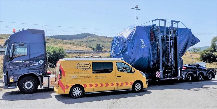 A large blue tarpaulin-covered load on a flatbed truck alongside a smaller van. Convoi Pilot Car with Rolls Royce Engine